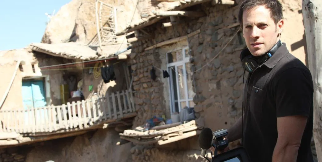 Adam Ellick stands in front of a row of houses, with headphones resting around his neck and a camera in his hand.
