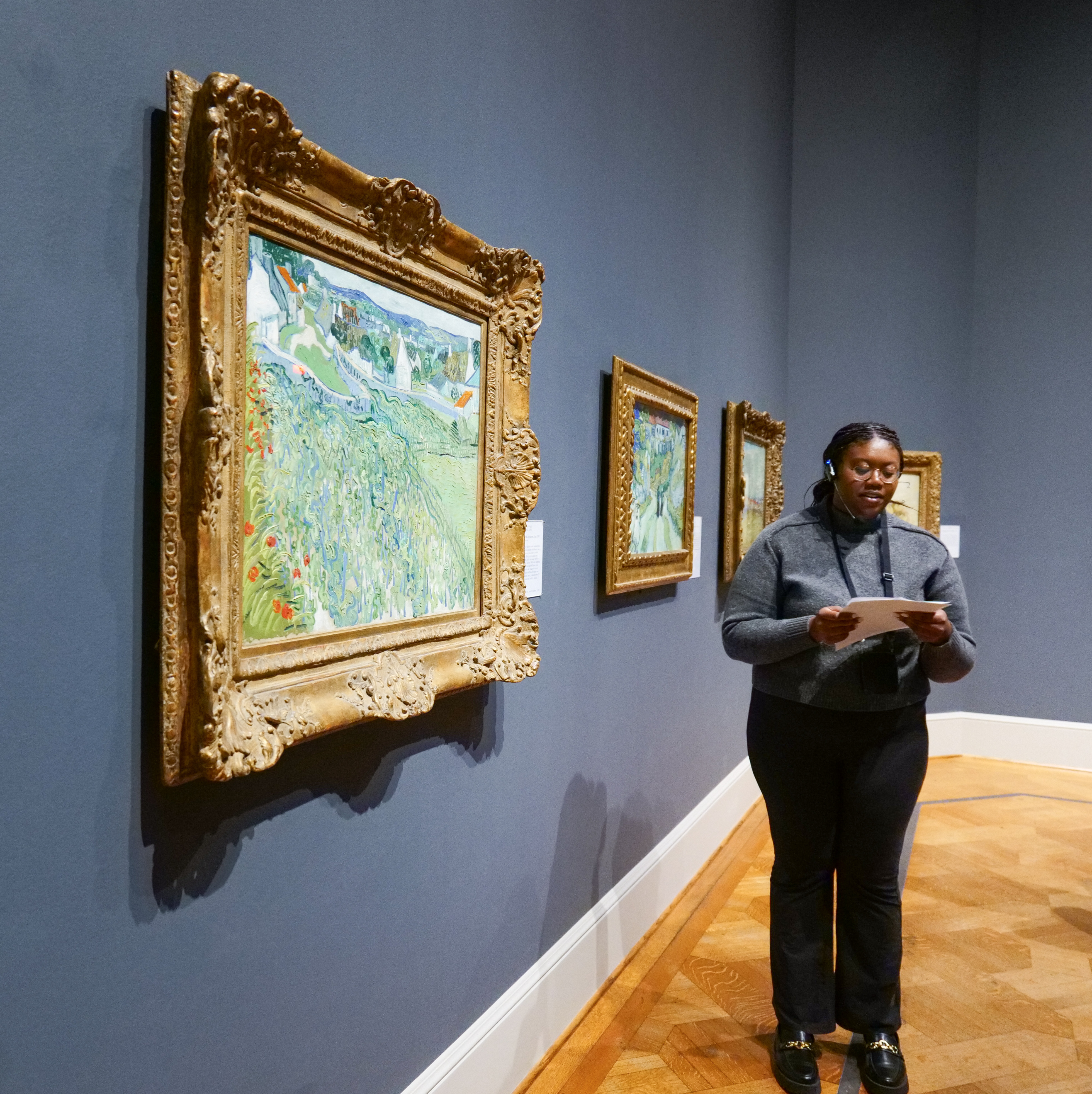 A woman standing next to a framed Van Gogh painting in a gallery. She has on a microphone and appears to be talking about the art.