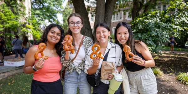 Four WashU College of Arts & Sciences students enjoying pretzels shaped like ampersands, the symbol of the college, while standing outside on campus. It's a sunny day during orientation, and the students are relaxed, laughing together.