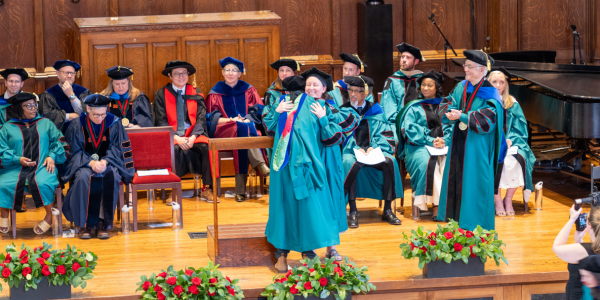 A faculty member and graduate student embrace on stage at the 2025 hooding ceremony.