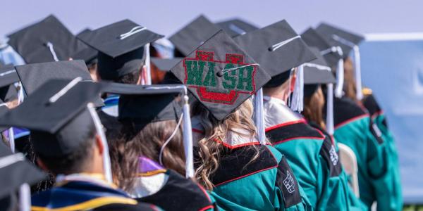 Students seated at the 2025 ceremony, with one graduation cap prominently featuring a WashU logo design.