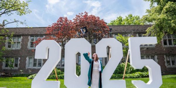A 2025 A&S graduate tosses their cap in front of a giant 2025 sign.