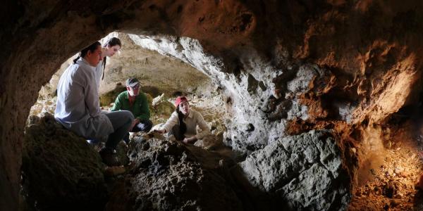 A group sits at the entrance of a cave in Sicily, peering into the cave and discussing its archaelogical significance.