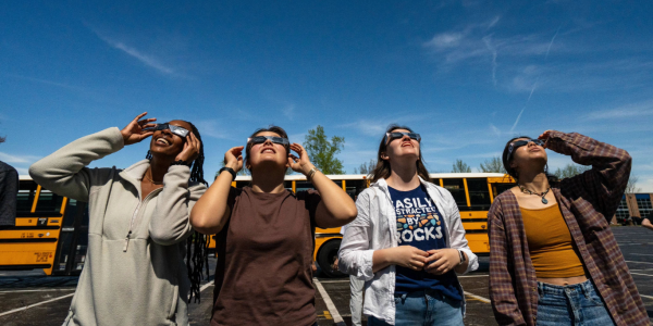 A group of WashU students standing in front of yellow school buses, all gazing up at the sky with eclipse glasses on. 