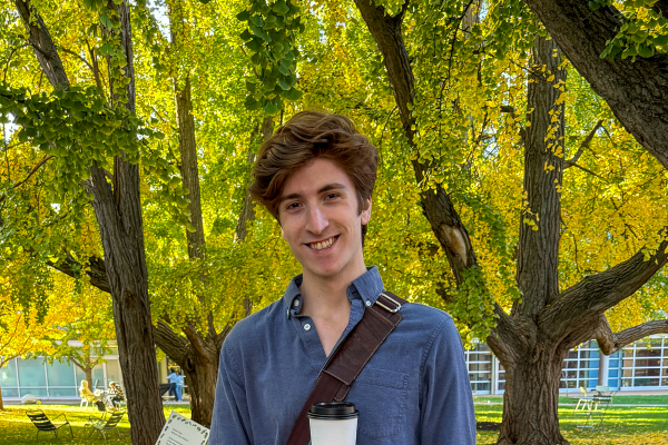 Student Zach Cohn, holding a coffee cup, smiles for a photo on a campus walkway.